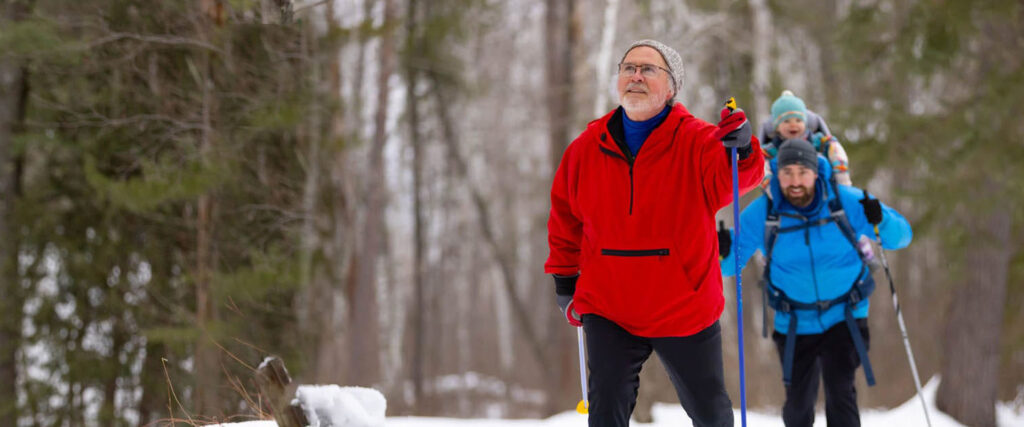 An older man cross country skiing in front of a younger man with his child strapped to his back.