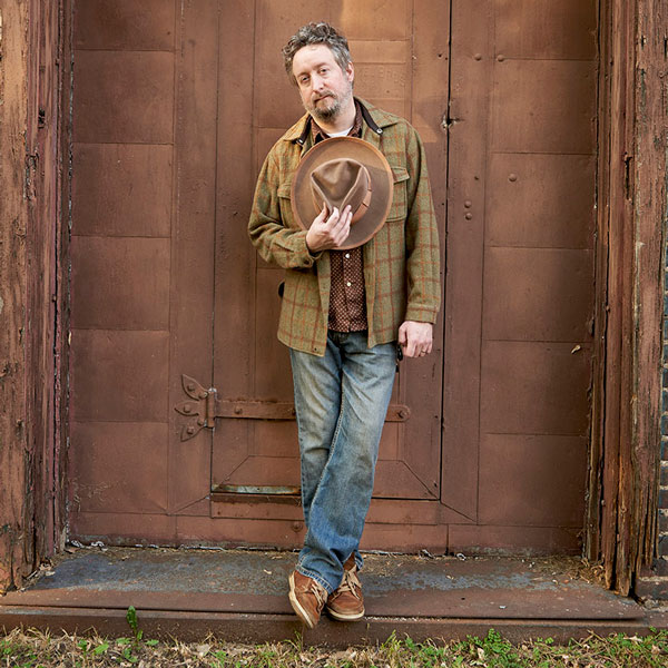 A man standing in front of an old brown door with a cowboy hat over his heart.