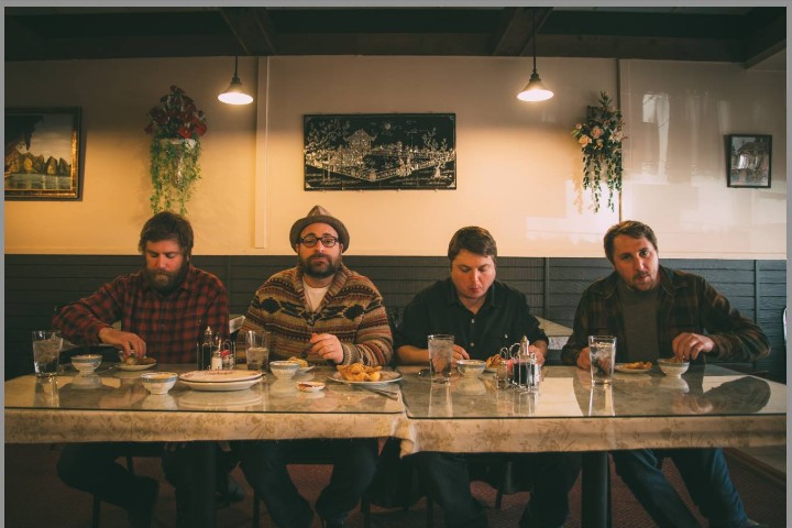 Four middle-aged men sitting at a table facing the camera eating a meal