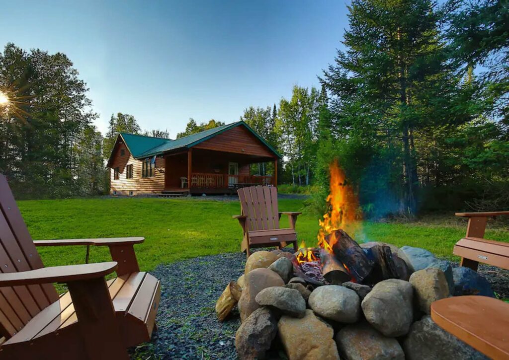 A calm campfire with wood chairs surrounding it with a log cabin in the background.