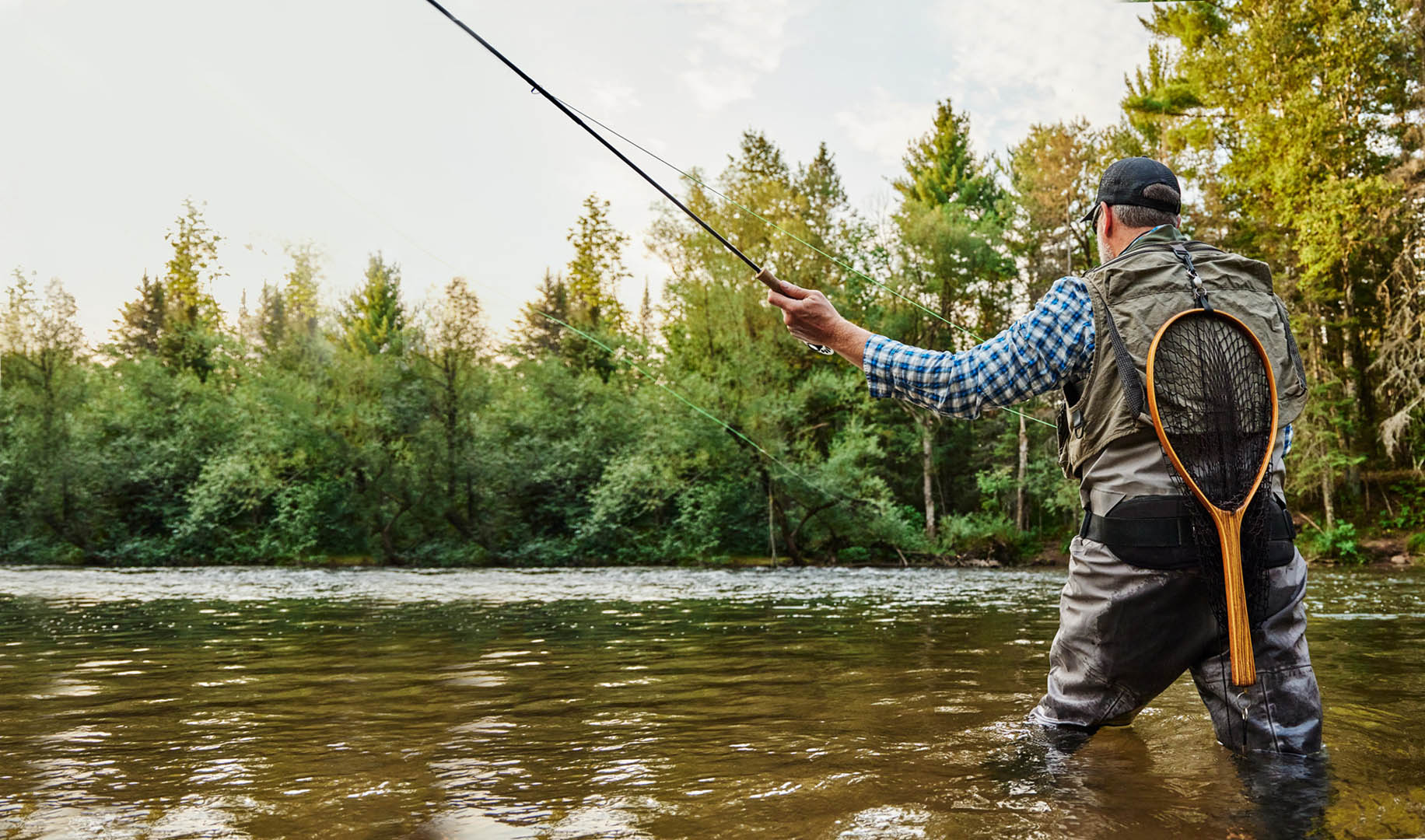 A man casting a fly fishing rod on a river surrounded by green trees.