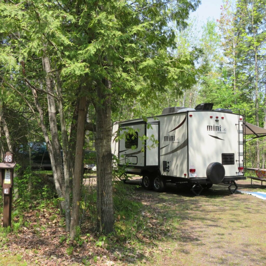 A fifth-wheel camper sits at a campsite surrounded by green trees.