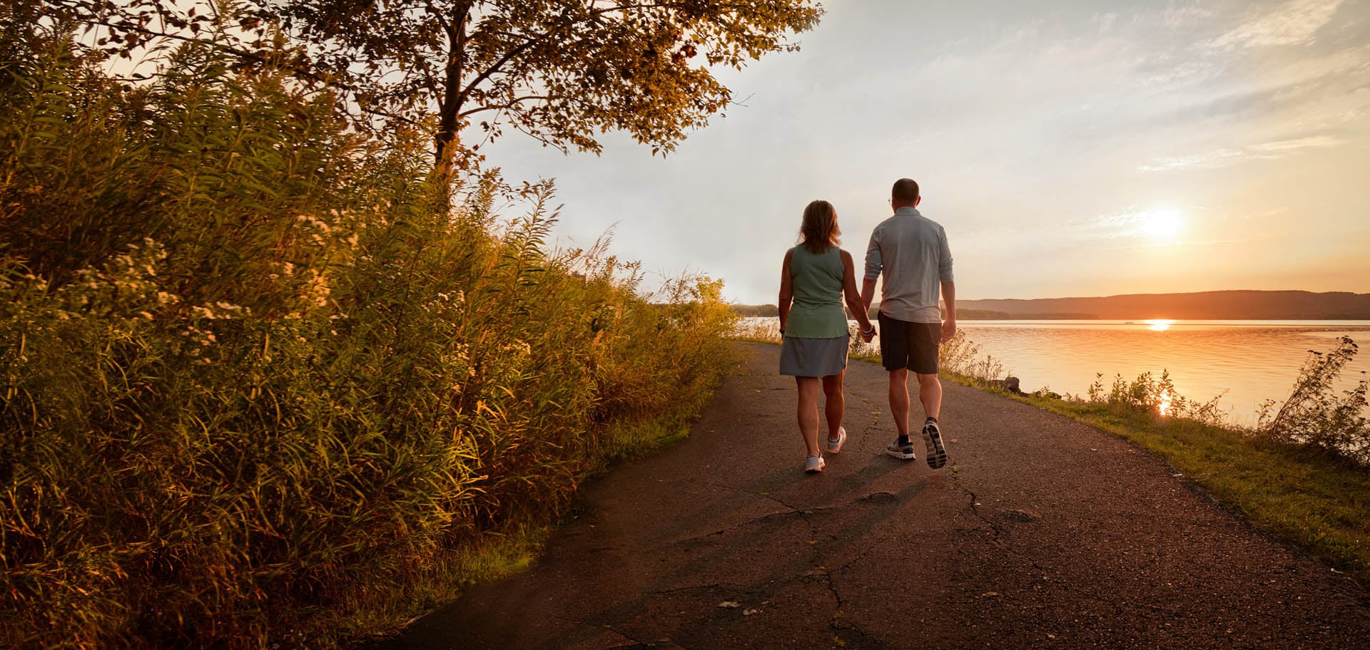 A couple holding hands as they walk down a path next to water at sunset.