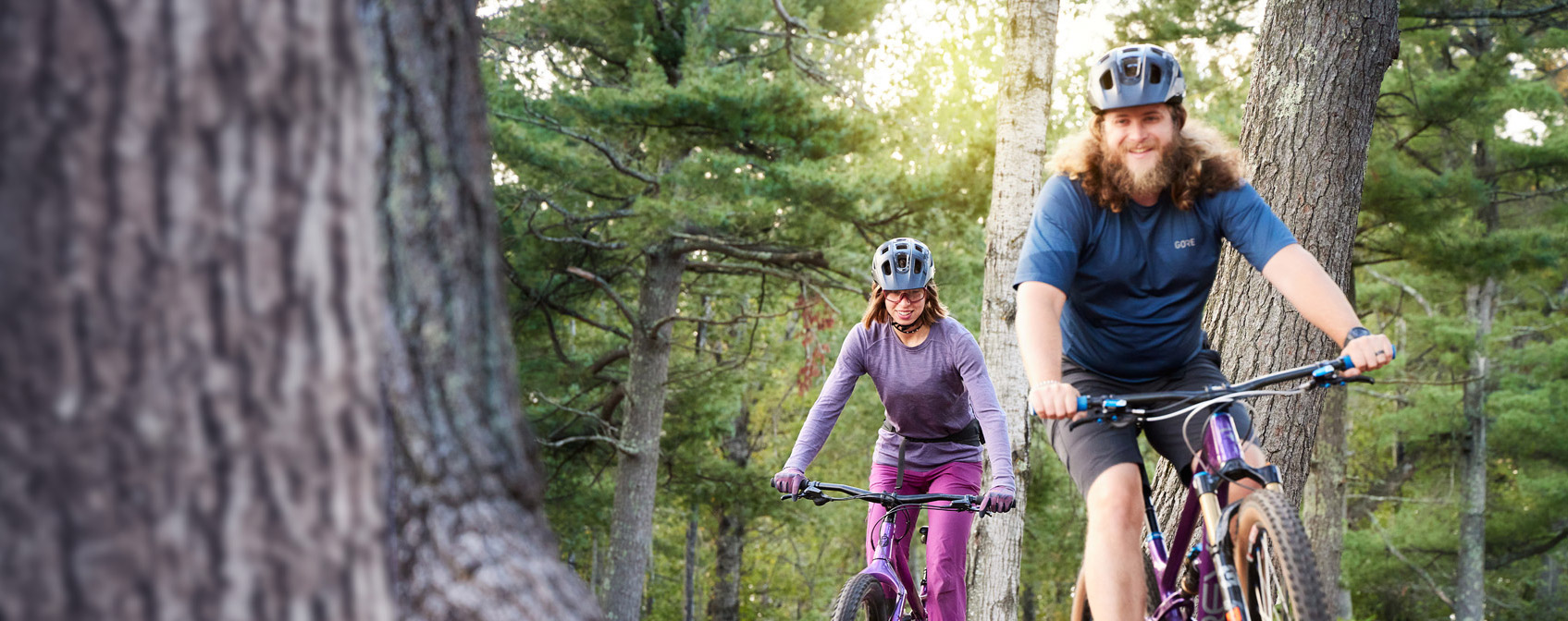 One male and one female mountain biker pedaling through the woods.