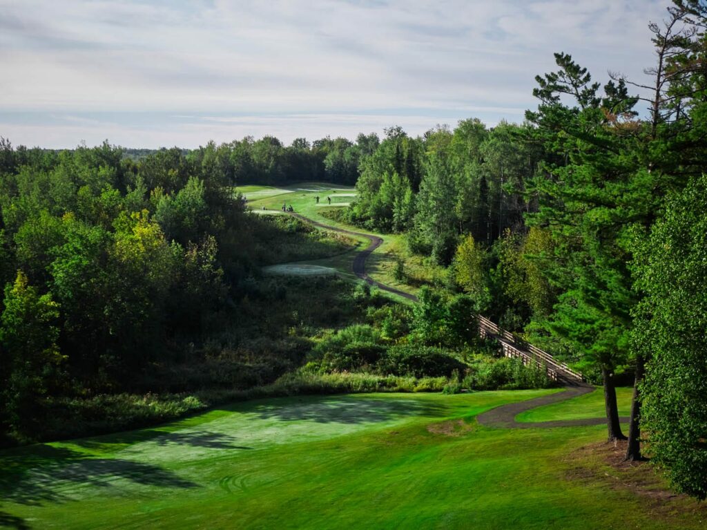 A scenic golf course in Superior, Wisconsin, with rolling green fairways, a winding cart path, and surrounding trees under a partly cloudy sky. A wooden bridge crosses a small gap in the trees—perfect for golf in Superior Wisconsin.