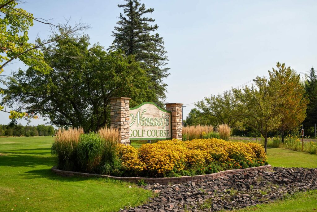 A large sign reading "Nemadji Golf Course" stands among yellow flowers and tall grasses, backed by stone pillars and surrounded by green trees—inviting you to enjoy golf in Superior, Wisconsin under a clear sky.