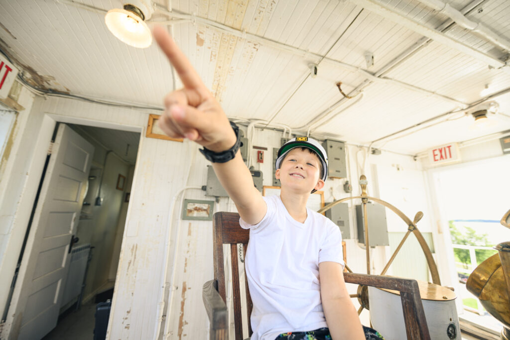 A young boy in a sailer cap pointing out the window of the SS Meteor. He's in the engine room with the large steering wheel behind him.