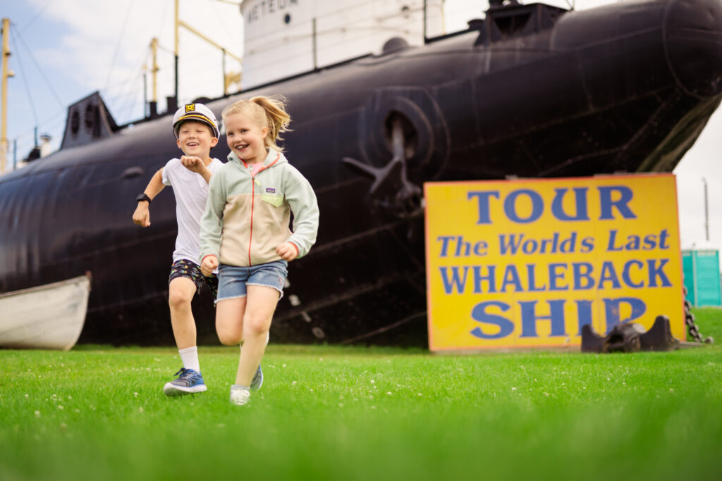 Two children running and laughing near a ship and its a sign that says "Tour the World's Last Whaleback Ship"