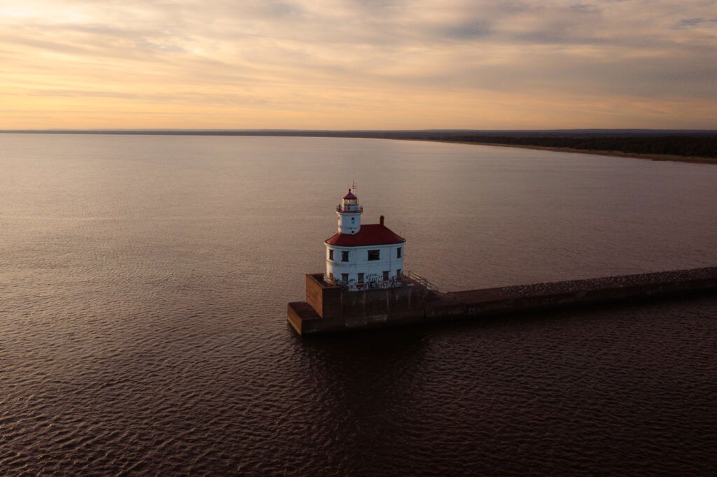 A lighthouse with a red roof sits at the end of a long pier, extending into a vast body of water. The sky is painted with warm hues of a sunset, and the horizon stretches into the distance.