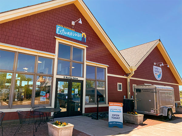 The image shows a red building with large windows, branded with "Lake Superior Estuarium" signage. A sandwich board outside reads "OPEN TODAY," next to a metal trailer. There are a few outdoor chairs and a clear blue sky overhead.