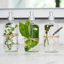 Three glass spray bottles are filled with water and different herbs. Each bottle is sealed with a silver cap and has twine wrapped around the midsection holding the herbs, which include thyme, mint, and lavender. They sit on a white surface.