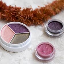 A makeup set featuring a round compact with three eyeshadow shades: pink, purple, and olive green. Two small jars of loose eyeshadow in mauve and dark purple are beside it on a textured surface, with a brown fluffy accessory nearby.