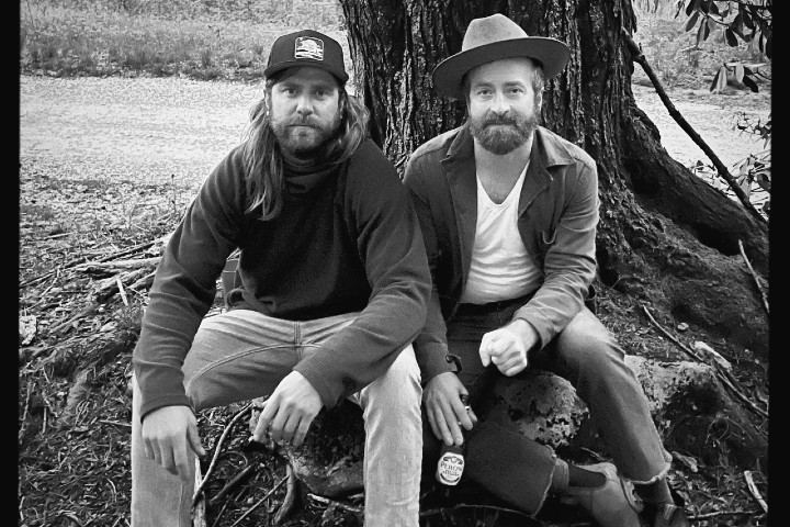 Two men sitting against a large tree trunk outdoors. One wears a cap and a dark jacket, while the other wears a hat and a light-colored jacket. They both have beards and are sitting on a rock with foliage around them. The image is in black and white.