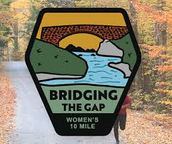 An autumnal forest forms the backdrop for a hexagonal badge with a bridge over a river and hills at sunrise. Text reads "Bridging the Gap Women's 10 Mile." A runner is faintly visible on the path.