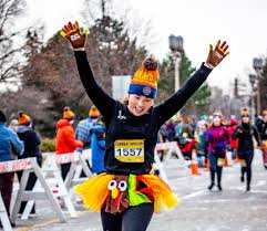 A person in a colorful turkey-themed costume crosses the finish line of a race, smiling with arms raised in victory. Other participants and spectators are blurred in the background. The atmosphere is festive.