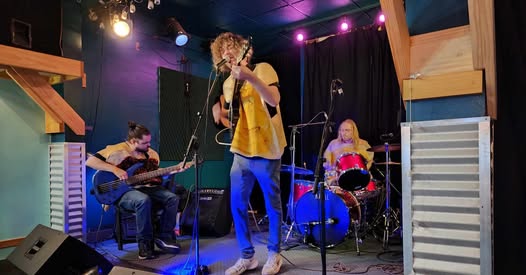 A band performs on a small stage. The guitarist and vocalist stands in the center, a bassist sits on the left, and a drummer plays on the right. The stage is decorated with colorful lights and wooden accents.