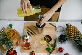 A person pours yellow liquid into a metal shaker on a wooden board surrounded by fresh ingredients including a pineapple, blueberries, herbs, a lemon slice, pomegranate seeds, and various utensils, suggesting a cocktail-making setup.