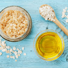 A glass jar of brown sugar, a wooden spoon with white salt, and a small glass bowl of golden oil are placed on a light blue wooden surface. Scattered sugar and salt granules decorate the table.