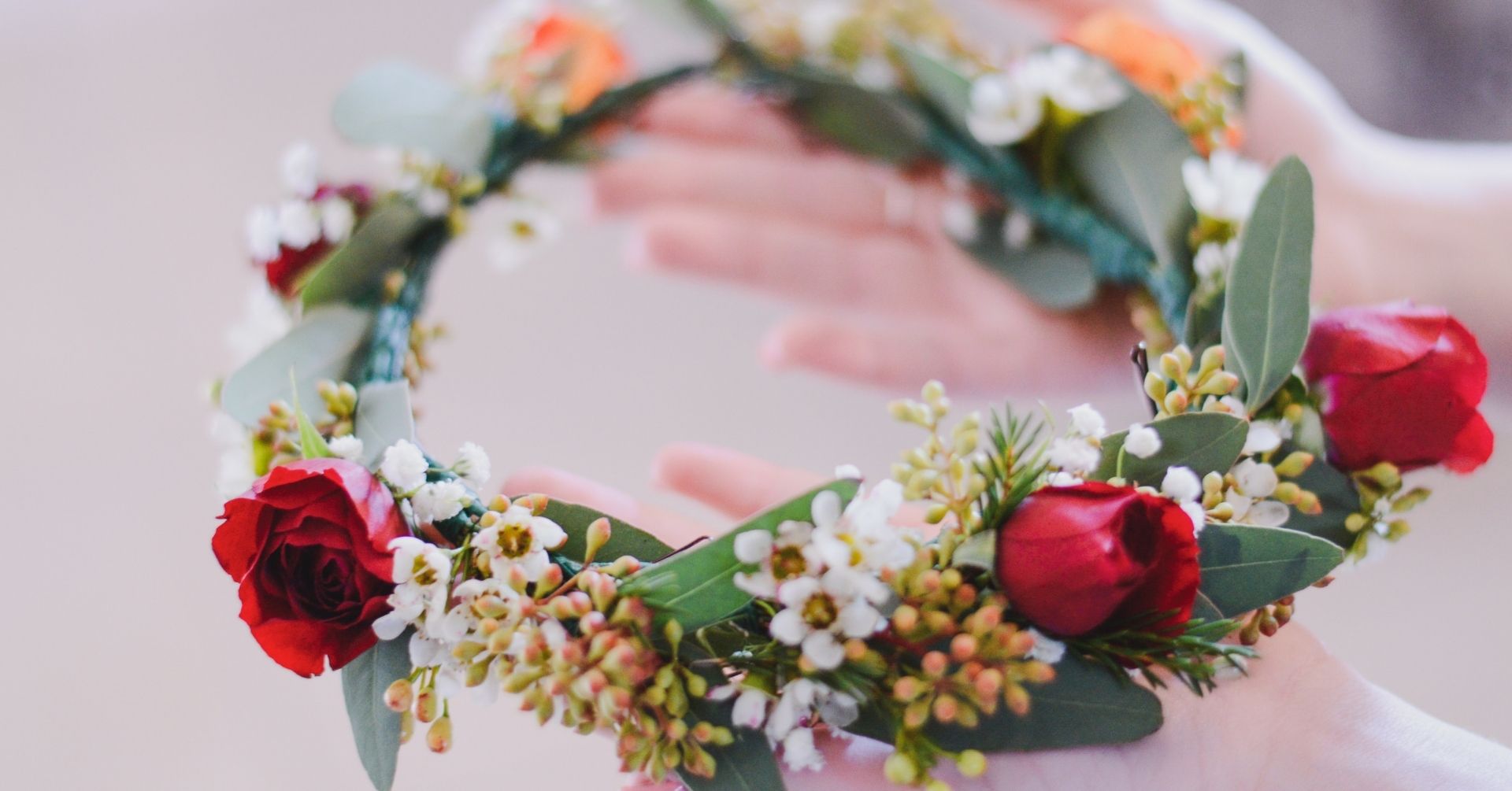 Hands holding a floral wreath adorned with red roses, small white flowers, and green leaves.