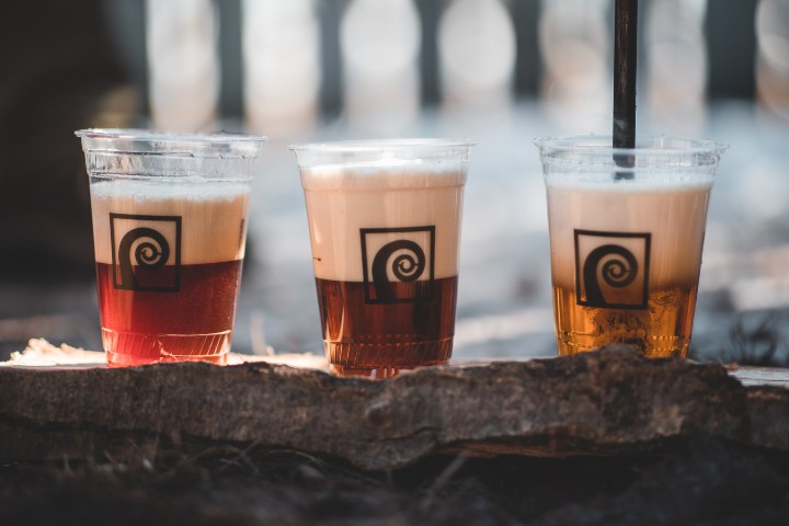 Three plastic cups filled with layered iced drinks sit on a wooden surface outdoors. Each cup displays a spiral logo. The beverages feature a distinct separation between frothy cream and darker liquid. Blurred background of vertical lines.