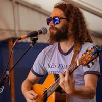 A person with long curly hair and sunglasses is playing an acoustic guitar and singing into a microphone. They are wearing a T-shirt and standing under a tent. The background is out of focus, showing a blue and white color scheme.