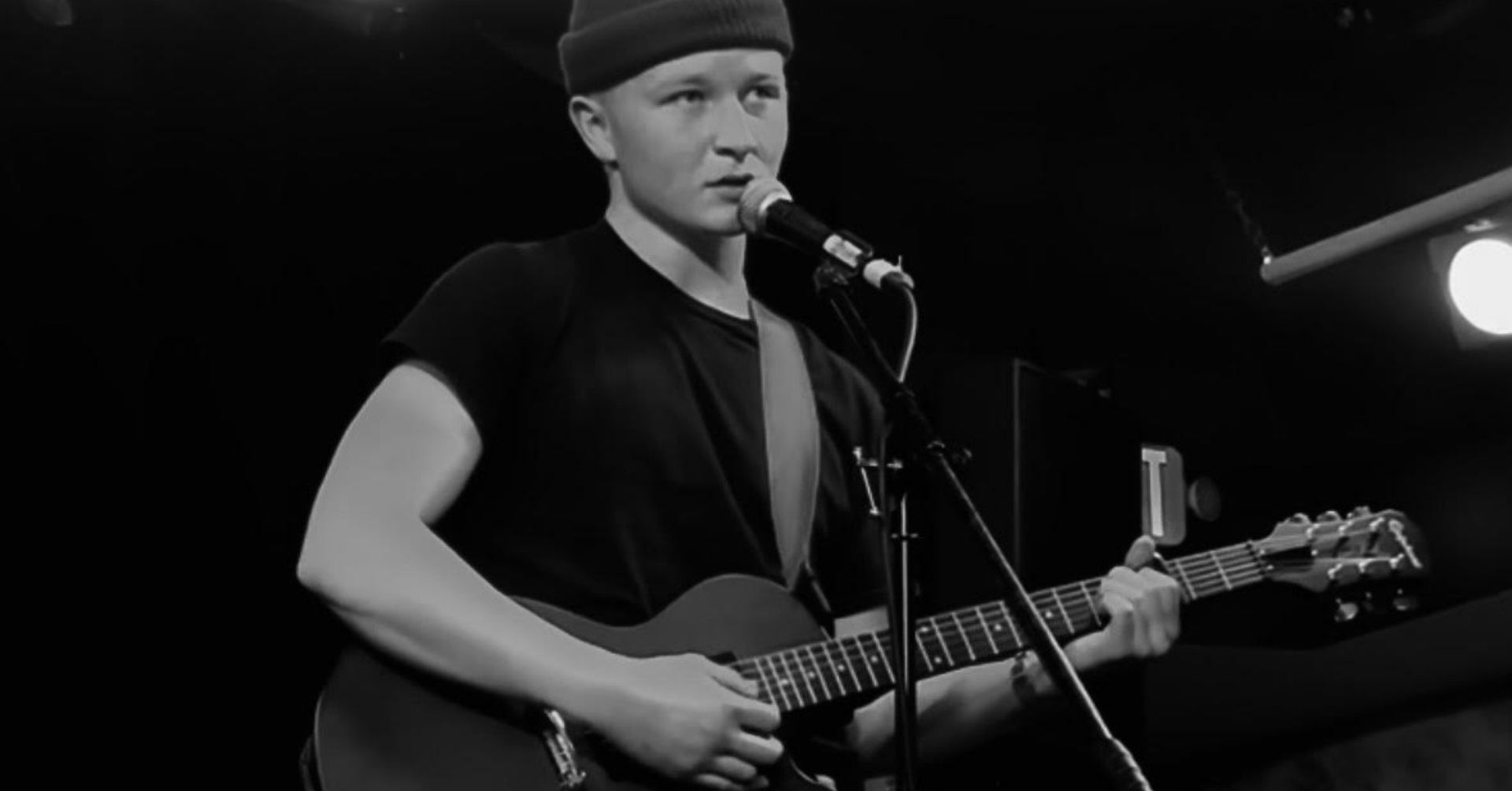 Person wearing a beanie and black shirt playing an acoustic guitar on stage, speaking or singing into a microphone. The background is dimly lit with stage lights.