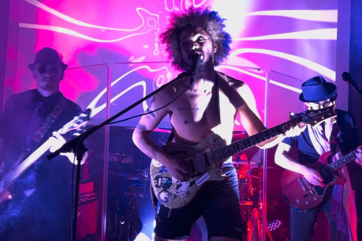 A shirtless guitarist with curly hair performs energetically on stage, illuminated by pink and purple lights. Two other musicians are in the background playing guitar. The atmosphere is vibrant and dynamic.