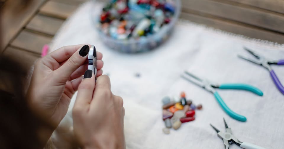 Close-up of hands using pliers to craft jewelry. Various colorful beads are scattered on a white cloth, with additional tools like pliers and a wire cutter nearby. The scene is set on a wooden table.