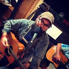 A musician with a beard and glasses plays the guitar and harmonica while singing into a microphone. Another person holding a guitar is partially visible in the background. The setting appears to be an intimate indoor space.