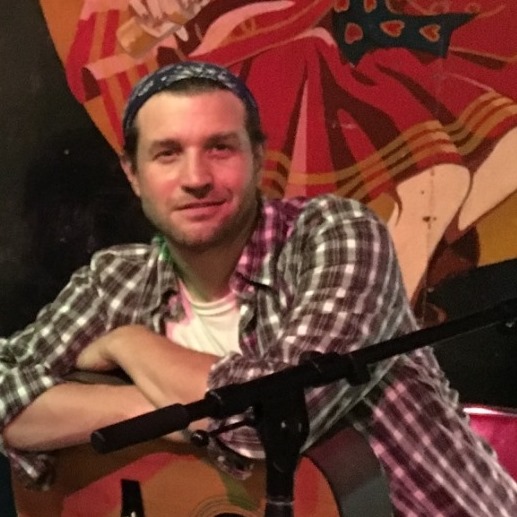 A person in a checkered shirt and blue bandana leans on a guitar with crossed arms, smiling. They are in front of a colorful mural, partly showing vibrant red and yellow designs. A microphone stand is visible in the foreground.