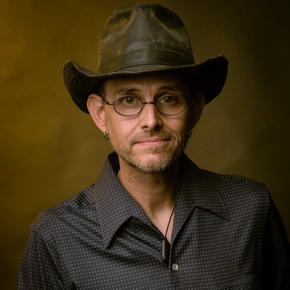 A man wearing a black hat, round glasses, a dark patterned shirt, and a necklace stands against a brown background, looking at the camera with a slight smile.