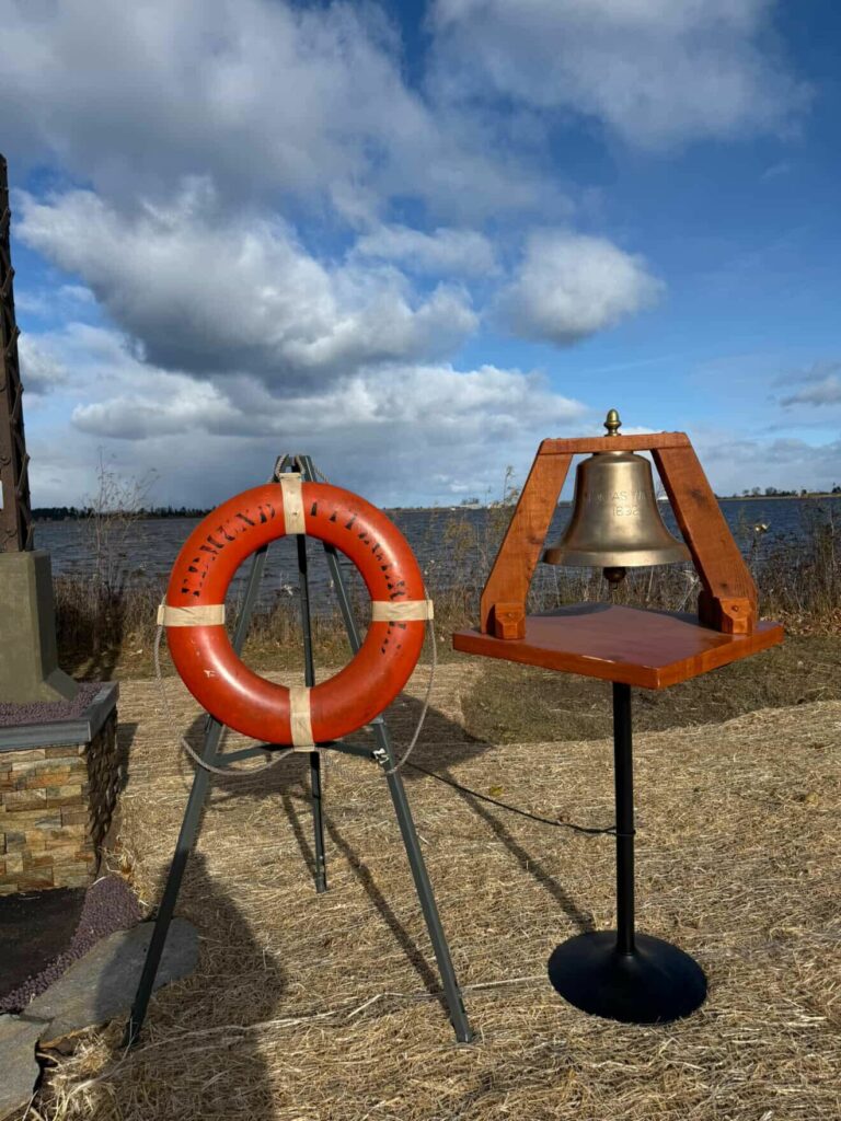 A brass bell on a wooden stand and an orange life preserver ring are displayed outdoors on dry grass, near a body of water under a partly cloudy sky.