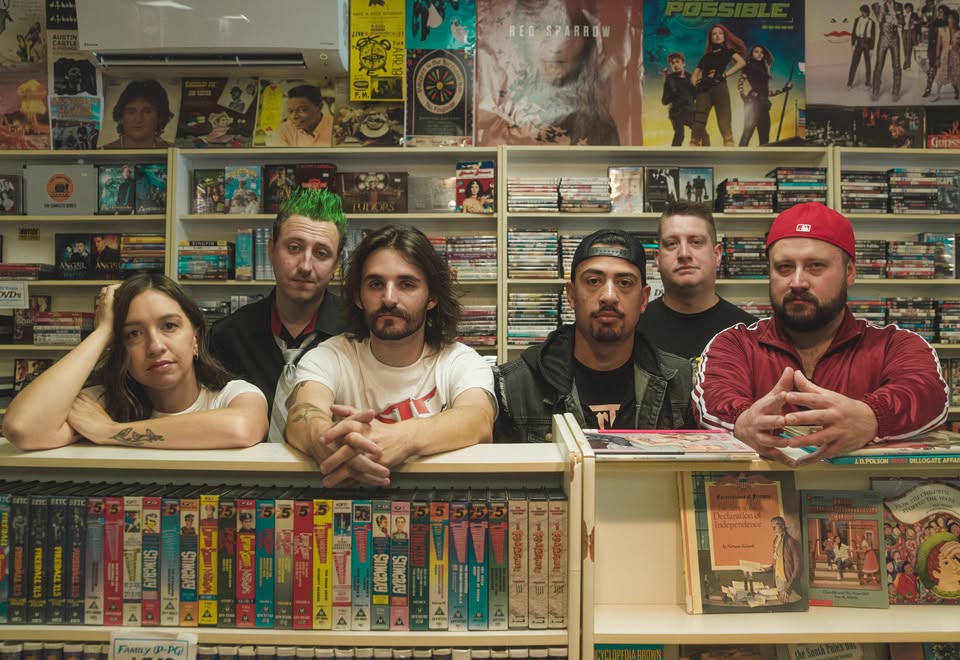 Six people pose behind a counter in a retro video rental store, surrounded by VHS tapes and movie posters on the walls, creating a nostalgic atmosphere.