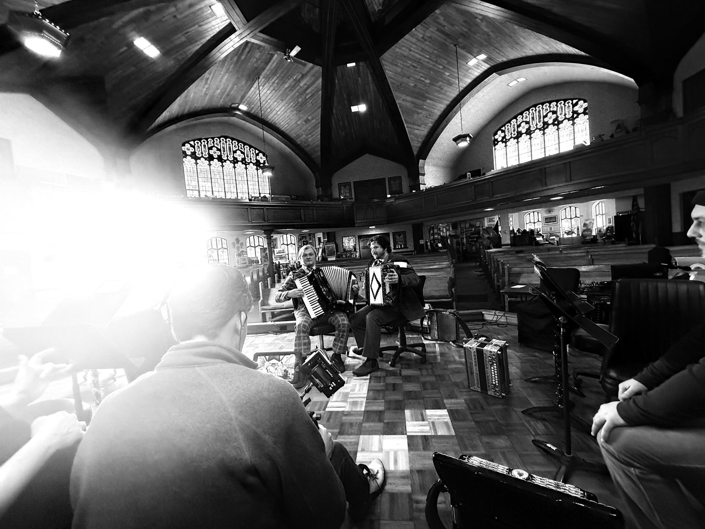 Black and white photo of musicians rehearsing in a church with vaulted wooden ceilings and stained glass windows. Two people play accordions while others sit nearby, some holding instruments. Bright light streams in from the left.