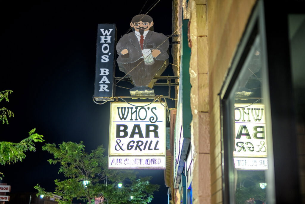 Neon signs for "WHO'S BAR & GRILL" and "WHO'S BAR" are mounted on a brick building at night, with a cartoon figure of a man in a suit holding a mug above the entrance. Trees and lights are visible in the background.