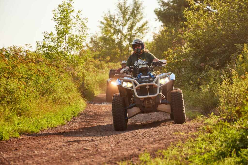 Two people enjoy summer activities in Superior, Wisconsin, riding ATVs on a dirt trail amid green bushes and trees. The lead rider, wearing a helmet and sunglasses, is followed by another ATV kicking up dust on a sunny day.