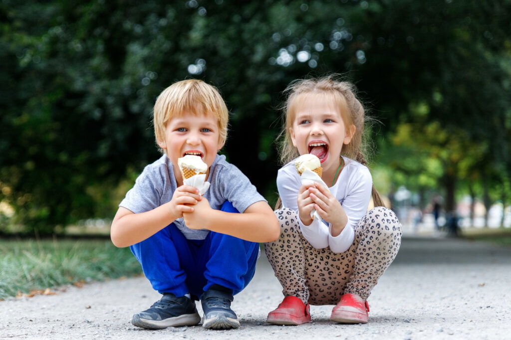 Two young children, a boy and a girl, crouch on a path outdoors, happily eating ice cream cones—one of the many delightful summer activities in Superior, Wisconsin. Trees line the background as they enjoy this sweet treat under the summer sun.