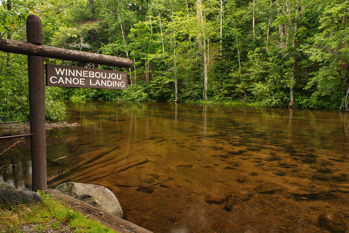 A wooden sign reading "Winneboujou Canoe Landing" stands by a calm, shallow river surrounded by lush green trees—an inviting spot for summer activities in Superior, Wisconsin. The clear water reveals rocks on the riverbed.