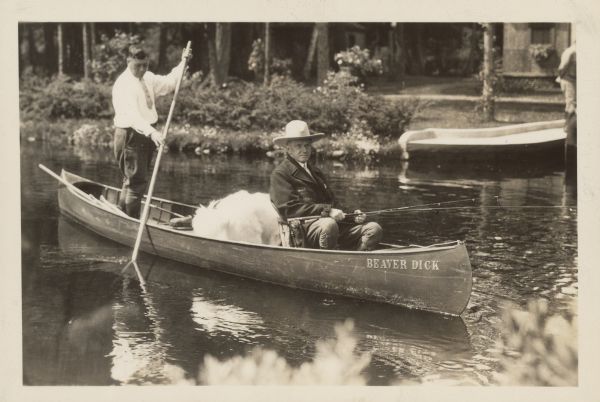 Two men in a canoe labeled "BEAVER DICK" enjoy summer activities in Superior, Wisconsin; one paddles while the other, wearing a hat, fishes. Trees and a docked boat are visible in the background on the calm water.