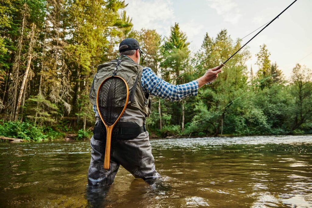A person wearing waders and a plaid shirt stands in a shallow river, fly fishing with a rod—one of the classic summer activities in Superior, Wisconsin. They are surrounded by trees and greenery, with a fishing net hanging on their back.