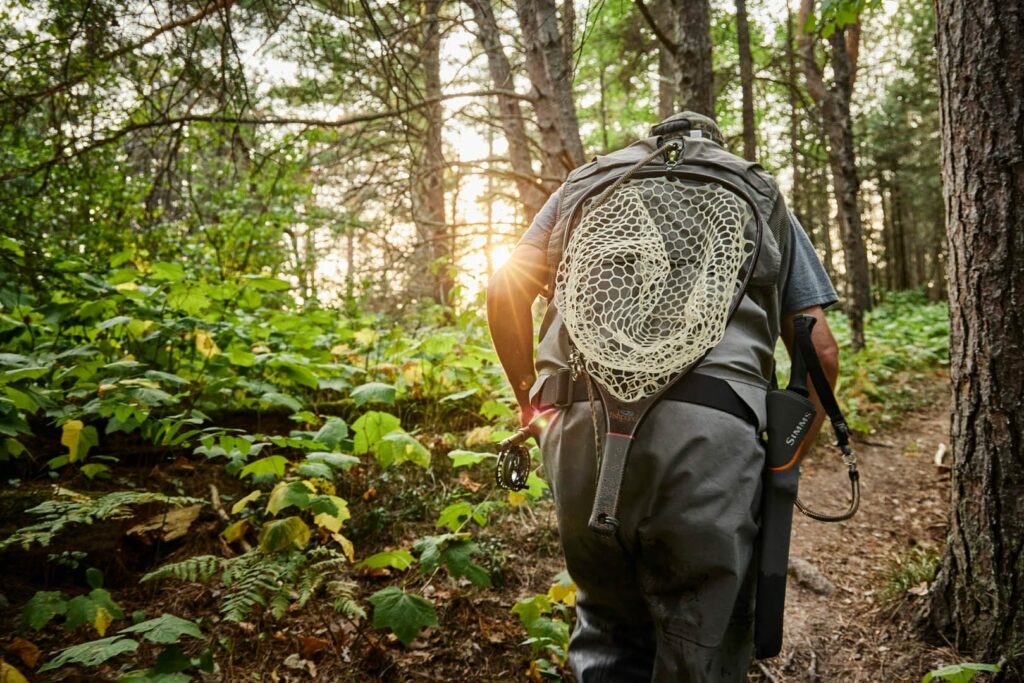 A person with fishing gear and a net attached to their backpack hikes through a sunlit forest trail, surrounded by green foliage and tall trees—one of the many summer activities in Superior Wisconsin.