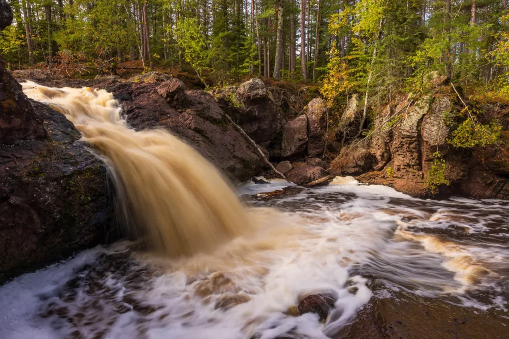 A small, powerful waterfall cascades over dark rocks into a swirling pool, surrounded by green trees—an ideal spot for summer activities in Superior, Wisconsin, where forest adventures await in the daylight.