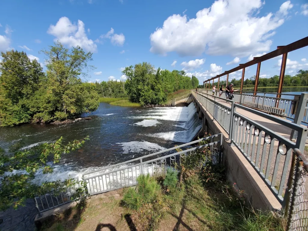 A riverside dam with water flowing over the edge, bordered by lush green trees under a bright blue sky. A walkway with railings runs beside the dam where a few people are standing, enjoying summer activities in Superior Wisconsin.