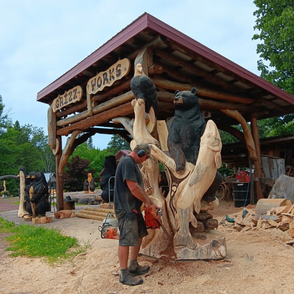 A man uses a chainsaw to carve a large wooden sculpture featuring animals, including a bear, eagle, and fish, outside "Grizz Works." Wood shavings cover the ground—one of the unique summer activities in Superior Wisconsin.