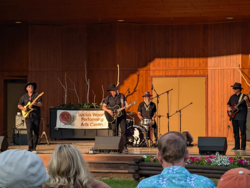 A four-member band dressed in black performs on an outdoor wooden stage as people watch from the audience—a perfect example of summer activities in Superior, Wisconsin. A sign on stage reads “Lucius Woods Performing Arts Center.” Warm sunlight fills the scene.