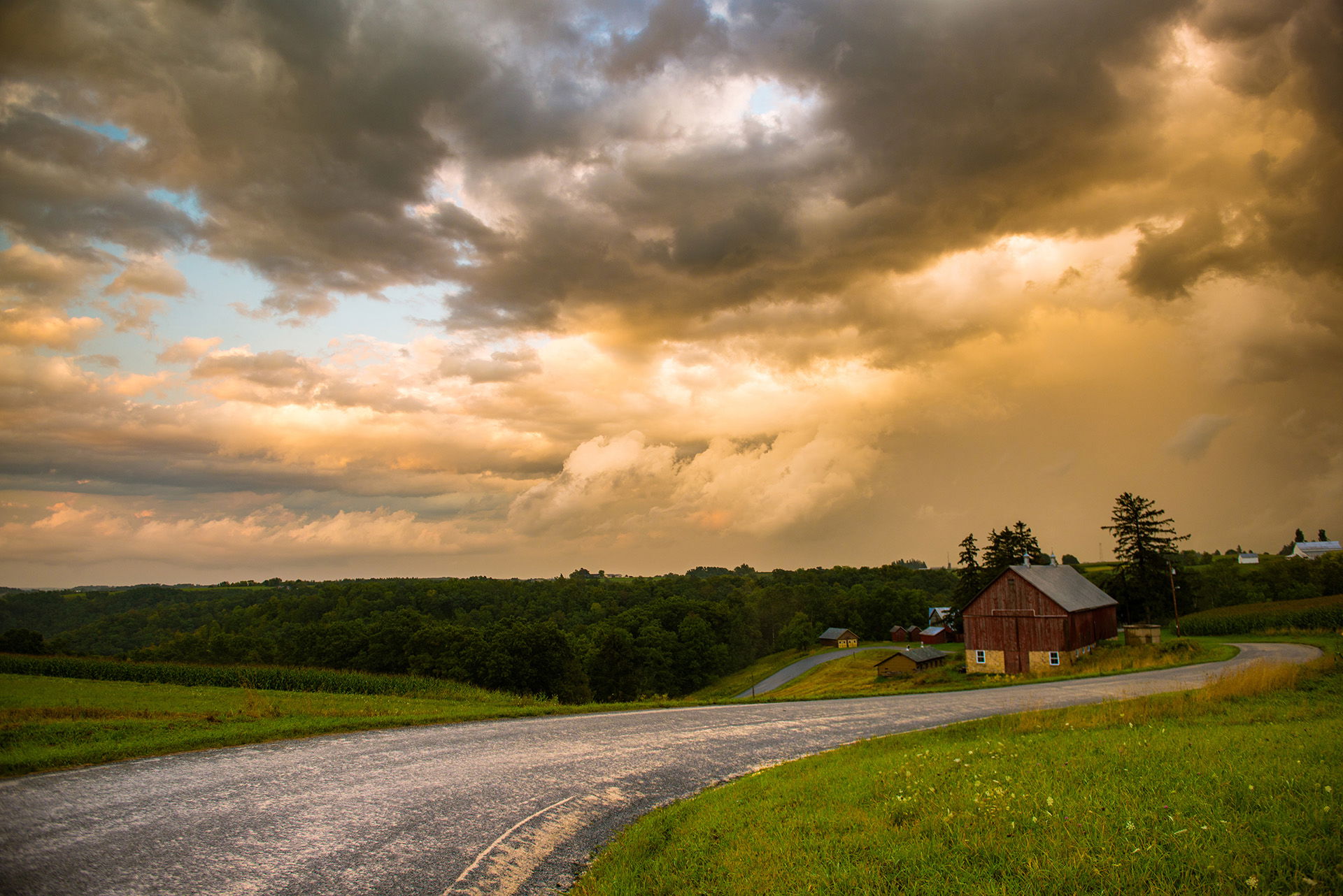 Shot of golden sky with clouds overlooking a barn and woods along a dirt road.