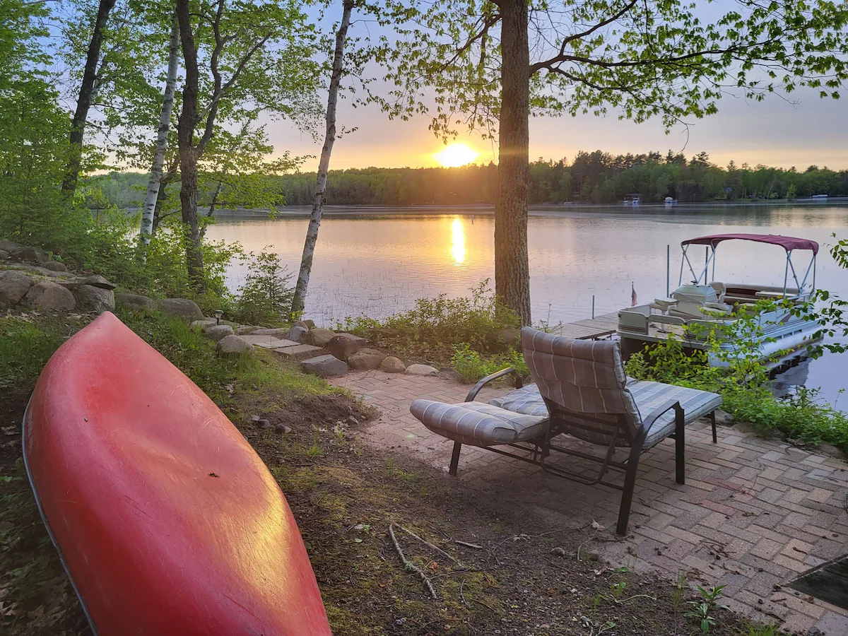 A peaceful lakeside scene at sunset in Superior, Wisconsin, with a red kayak on the ground—perfect for summer activities. Cushioned chairs on a brick patio, a dock with a boat, and trees reflecting in the calm water complete the view.