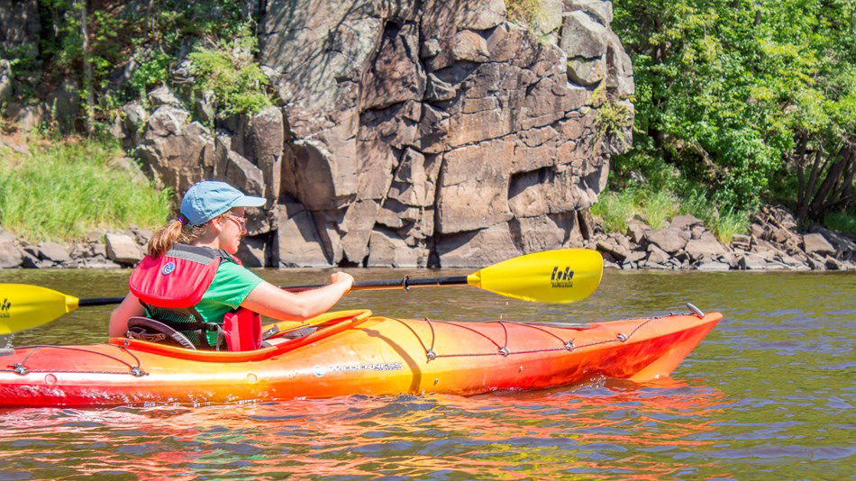 A person wearing a life jacket and blue cap paddles an orange kayak on a river, surrounded by rocky cliffs and green trees—enjoying one of the many summer activities in Superior, Wisconsin.