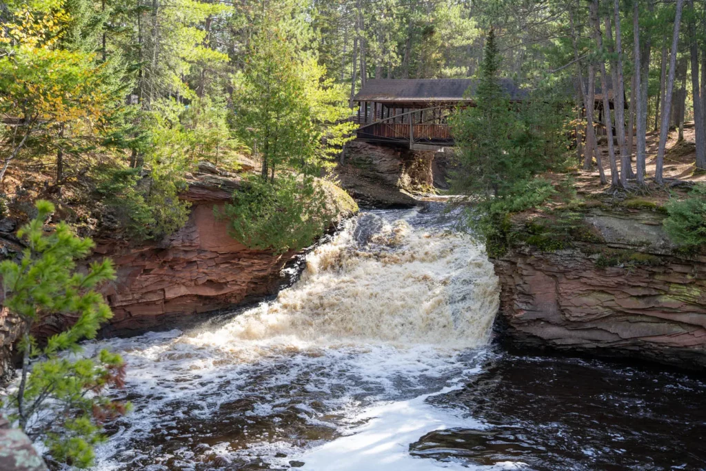A waterfall flows over rocky cliffs surrounded by green trees, with a covered wooden bridge spanning the stream above—a peaceful forest setting perfect for summer activities in Superior, Wisconsin.