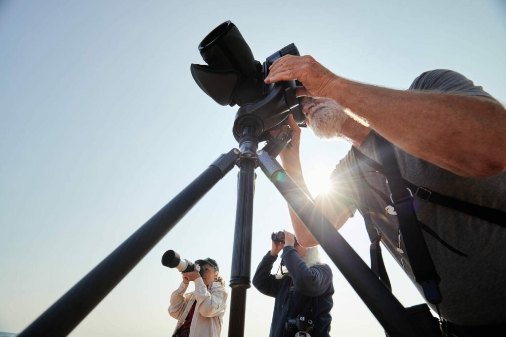 Three people enjoy summer activities in Superior, Wisconsin, using cameras with long lenses on tripods outdoors under a clear sky. Sunlight shines through, partially silhouetting a person in the foreground adjusting a camera.